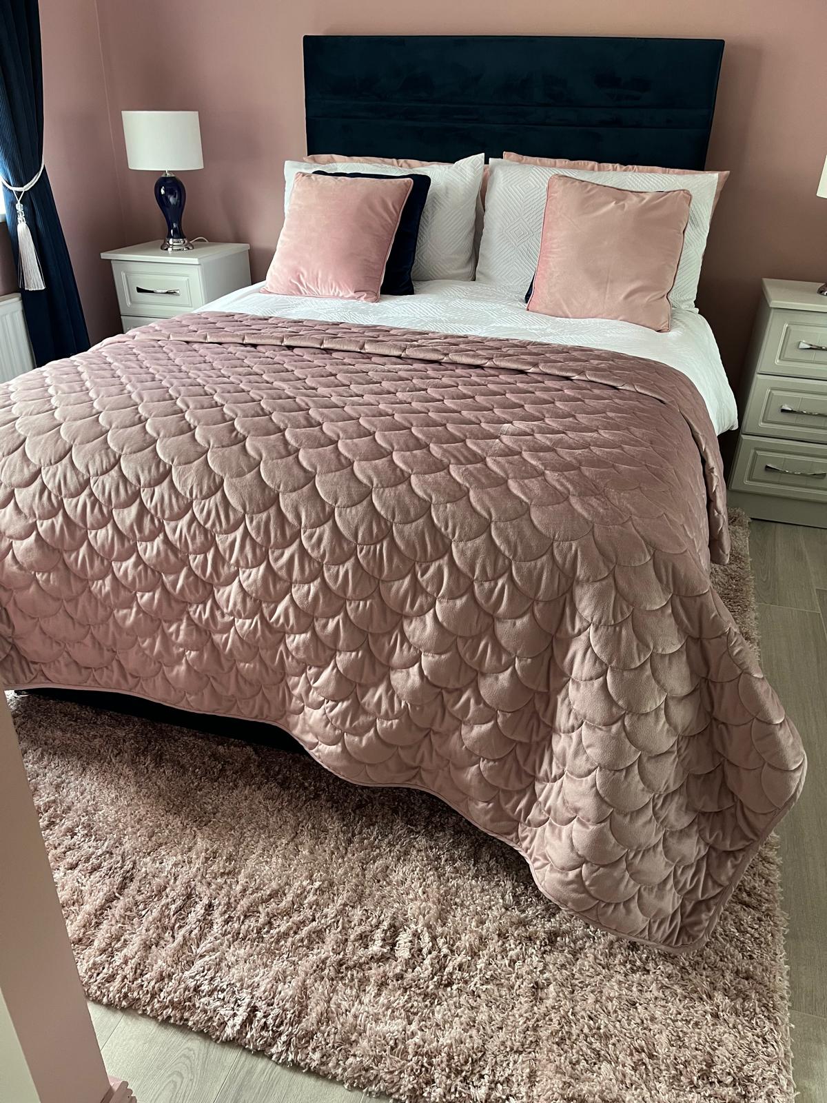 a bedroom with pink walls, a navy blue, velvet bed with two side tables. The side lamps have deep blue bases and white shades and are switched on. The bedclothes are white with a pink throw and there are two navy and two blush pink throw cushions. There is a shaggy blush pink rug under the bed.